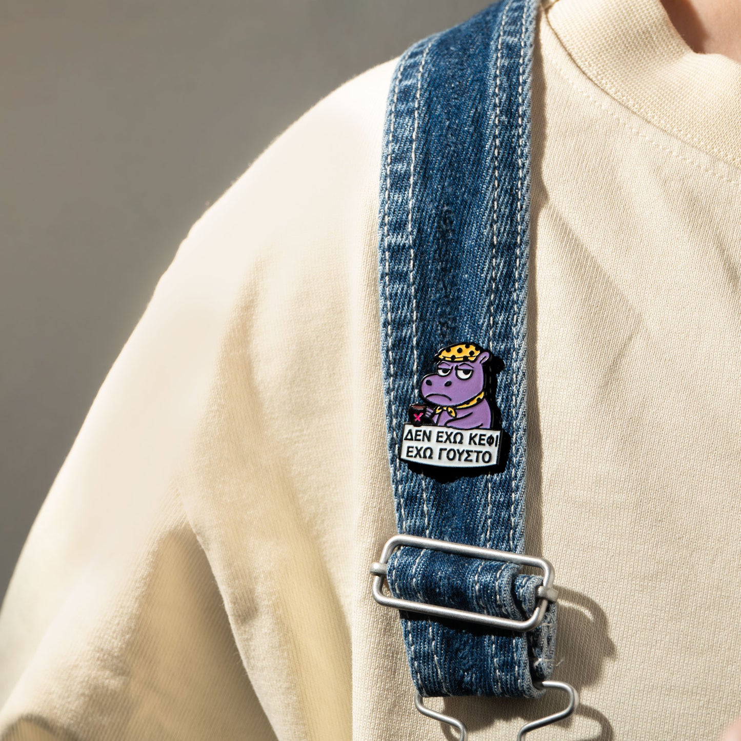 Denim strap with a cartoon hippo character enamel pin on it and a beige sweater