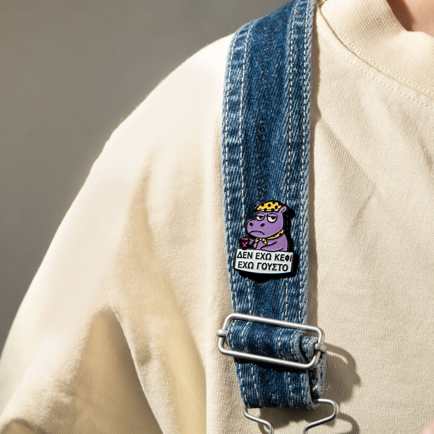 Denim strap with a cartoon hippo character enamel pin on it and a beige sweater